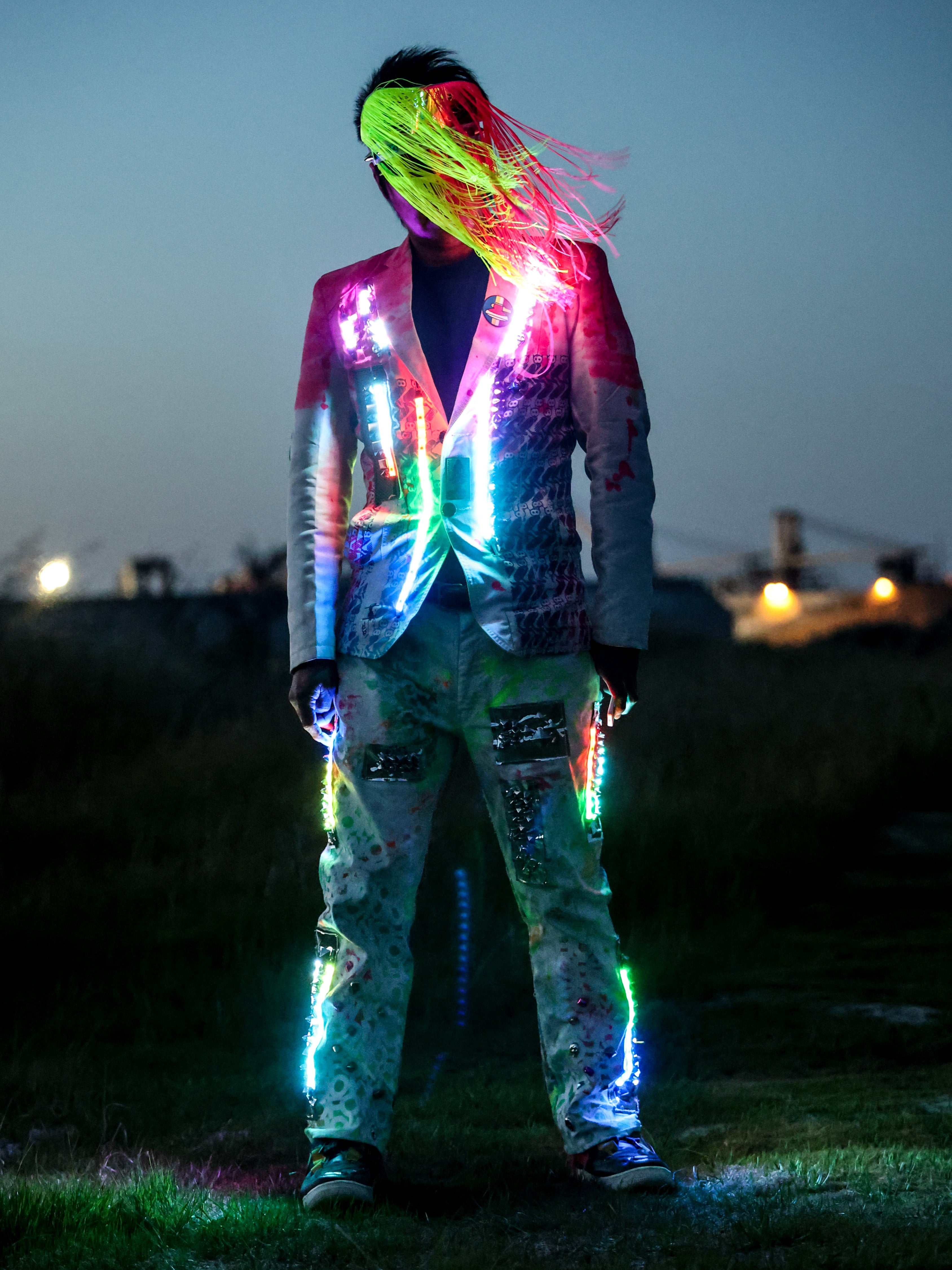 A person standing outdoors at dusk wearing a brightly lit, graffiti-style jacket and matching pants with glowing neon accents. The outfit reflects pink, green, blue, and purple lights against a dark evening sky and grassy background.