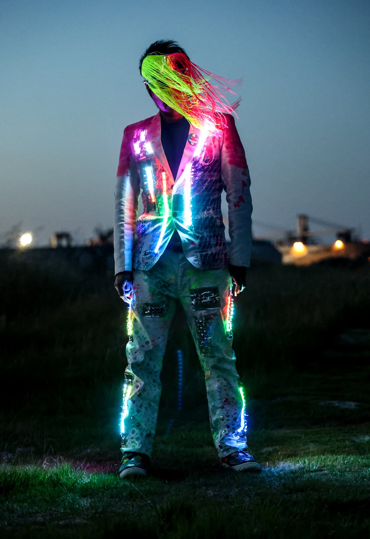 A person standing outdoors at dusk wearing a brightly lit, graffiti-style jacket and matching pants with glowing neon accents. The outfit reflects pink, green, blue, and purple lights against a dark evening sky and grassy background.